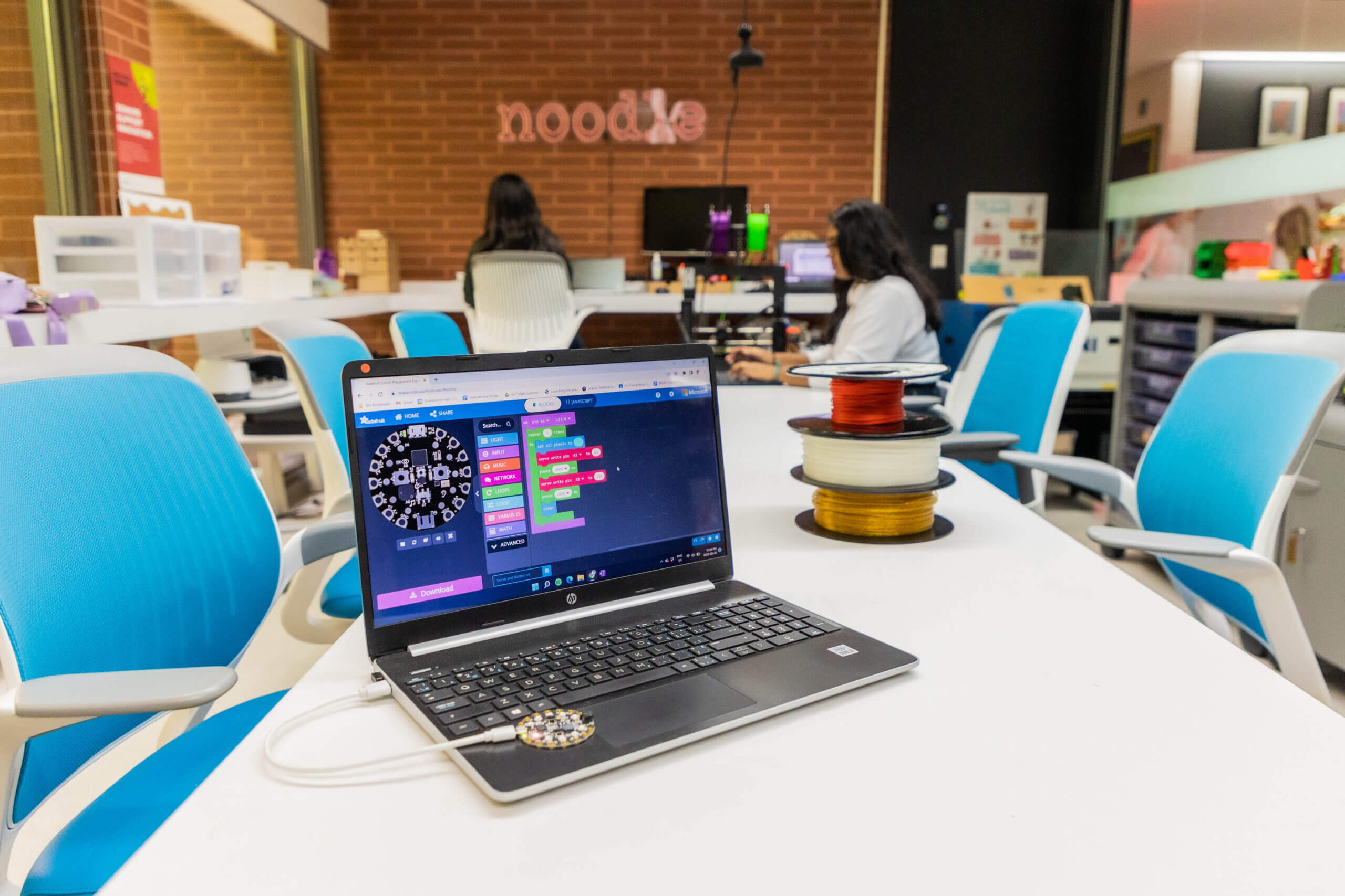 A laptop displaying a programming interface sits on a white table next to spools of colored filament in a modern, brightly lit workspace with blue chairs and people working in the background.