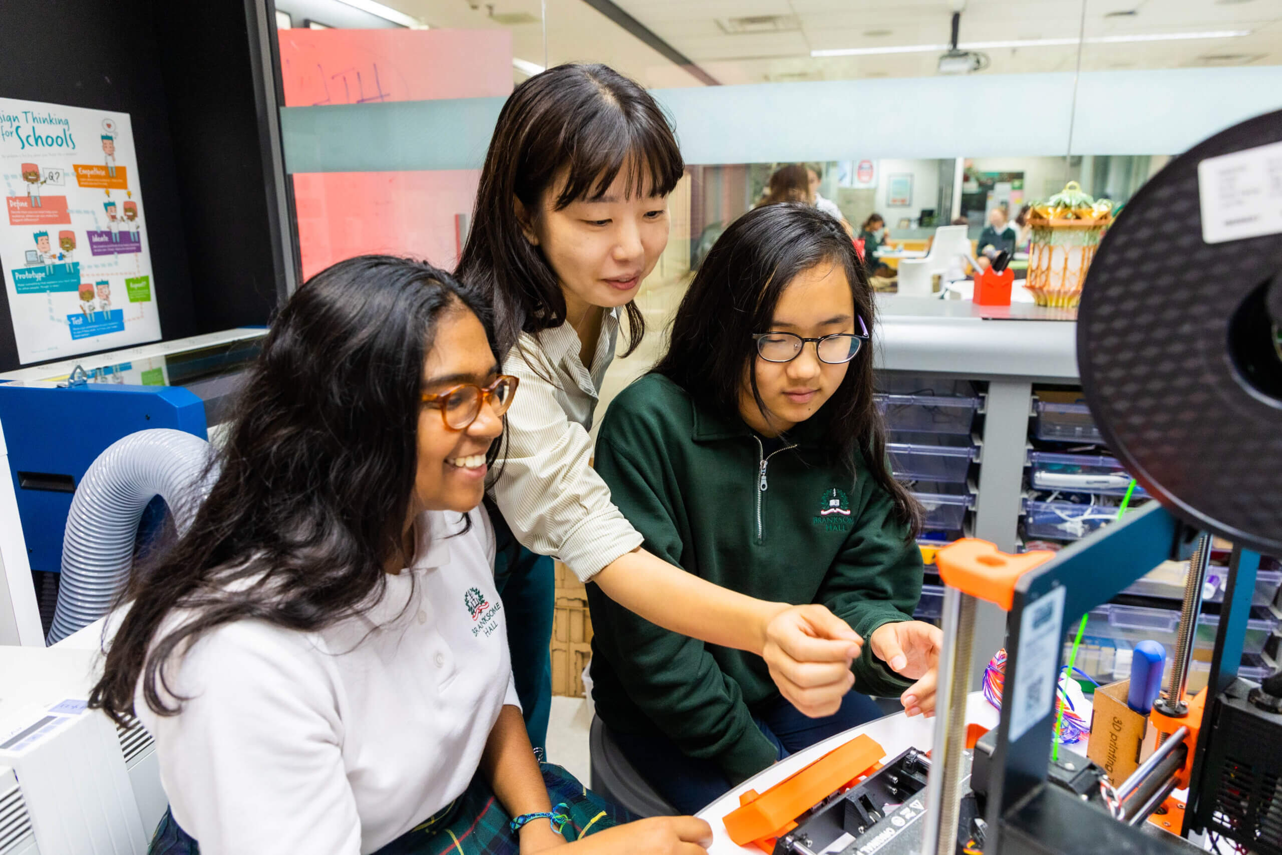 Three young women work together at a desk with a 3D printer in a bright classroom. One is guiding the others, who are focused on the printer. Shelves, tools, and colorful posters are visible in the background.
