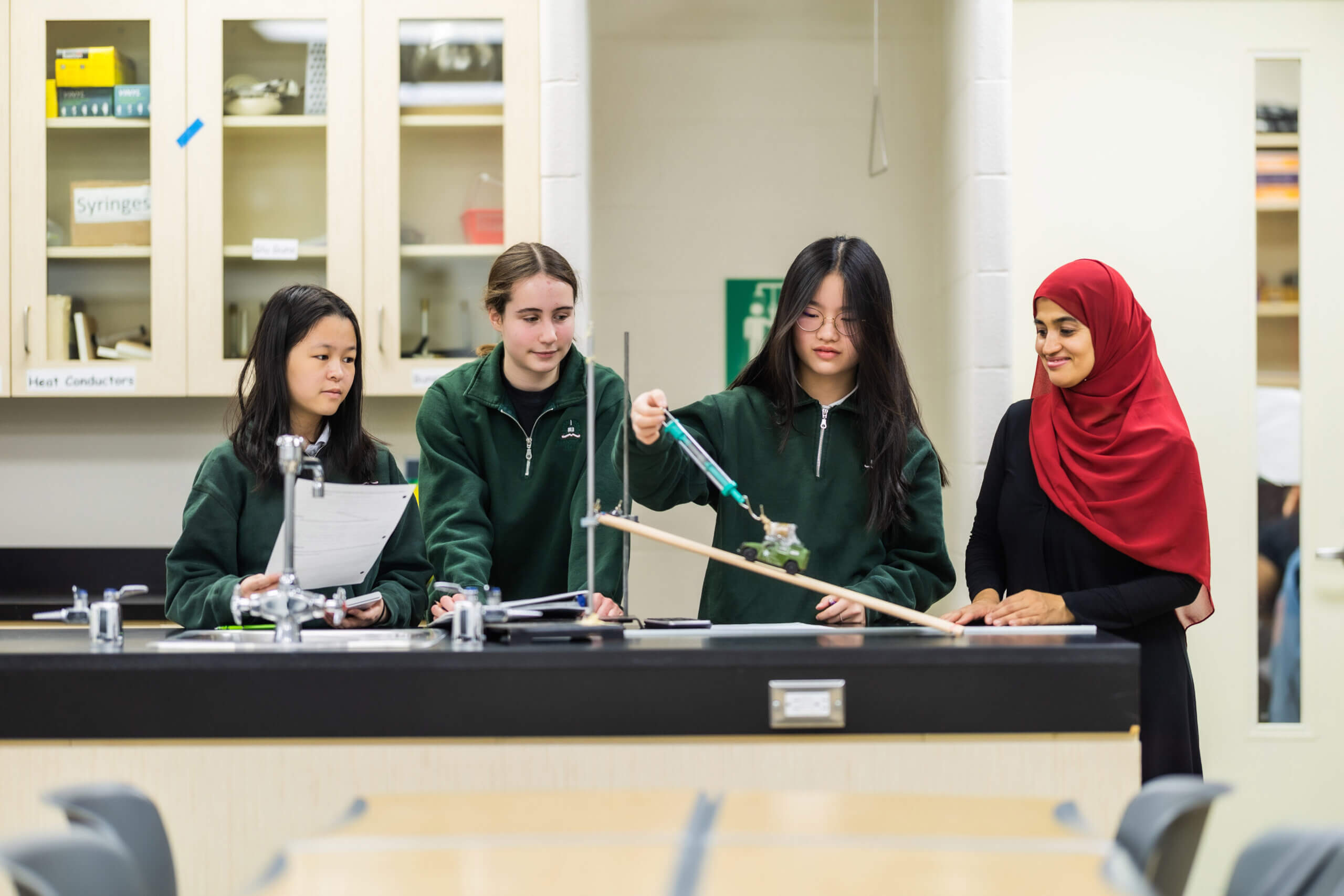 Four students in green uniforms and a teacher in a red hijab conduct a science experiment with a ramp and books in a classroom, observing results and taking notes.