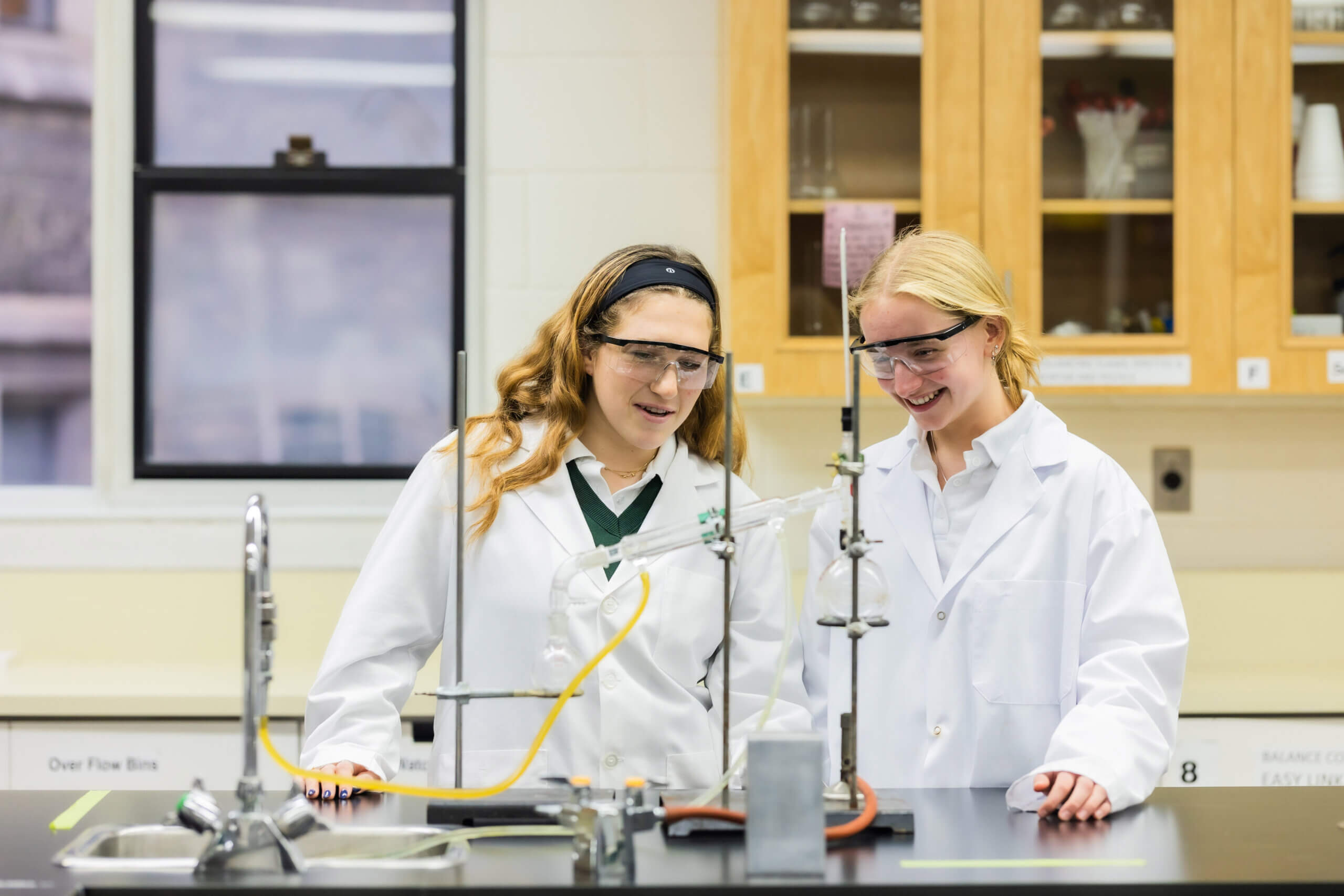 Two students in white lab coats and safety goggles stand at a lab bench, smiling while conducting an experiment with glassware and lab equipment in a bright science classroom.