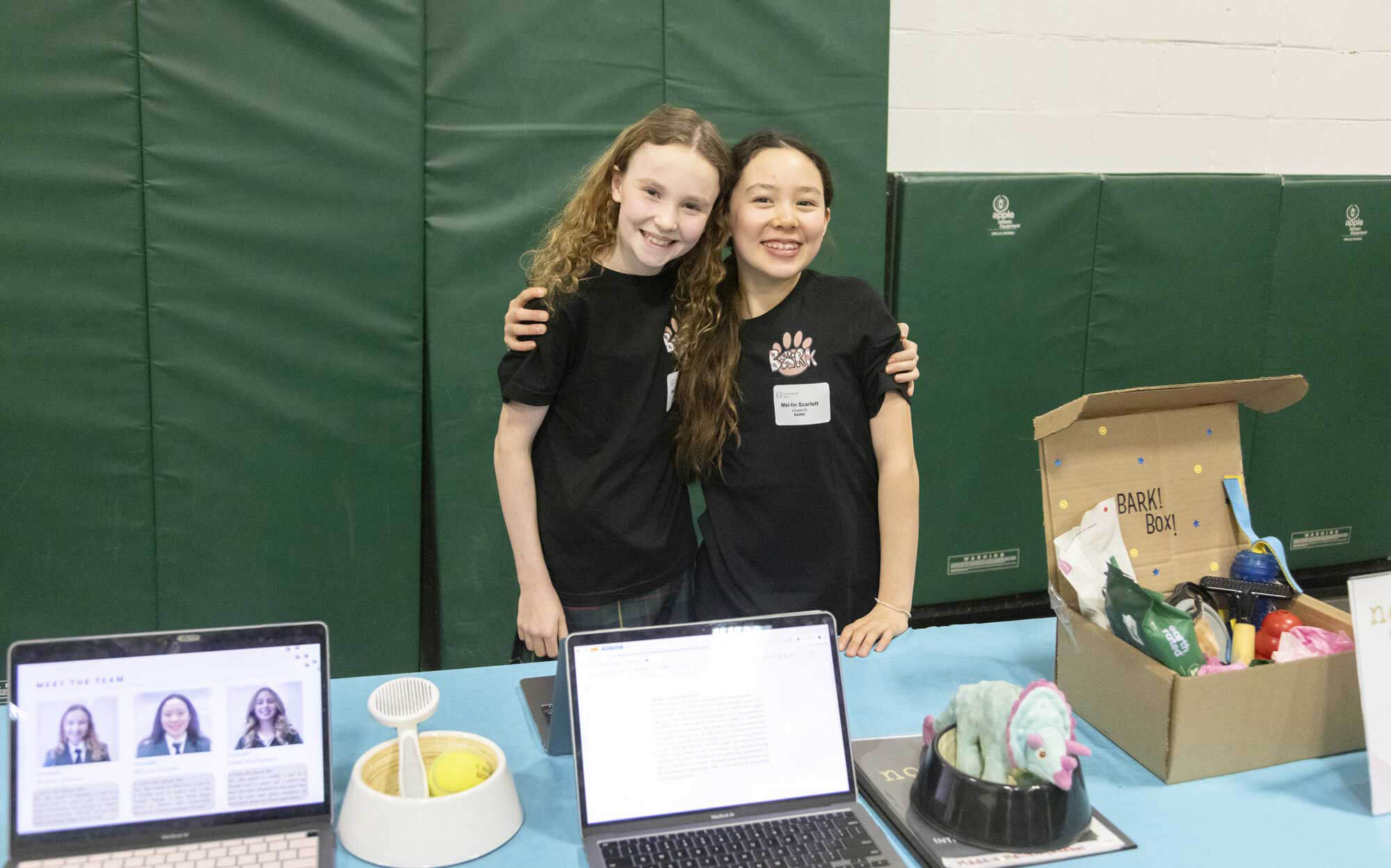 Two smiling girls stand behind a table with laptops, dog toys, and a BarkBox at what appears to be a school event or science fair. They are wearing black shirts and name tags, posing with arms around each other.