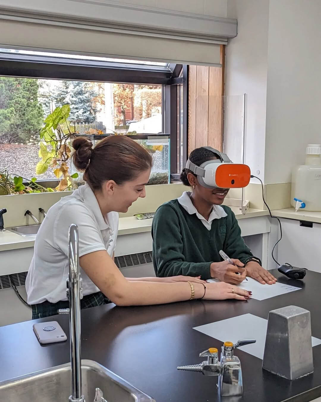 Two students sit at a lab table; one wears a virtual reality headset and writes on paper while the other smiles and observes. Sunlight comes through a window with plants in the background.