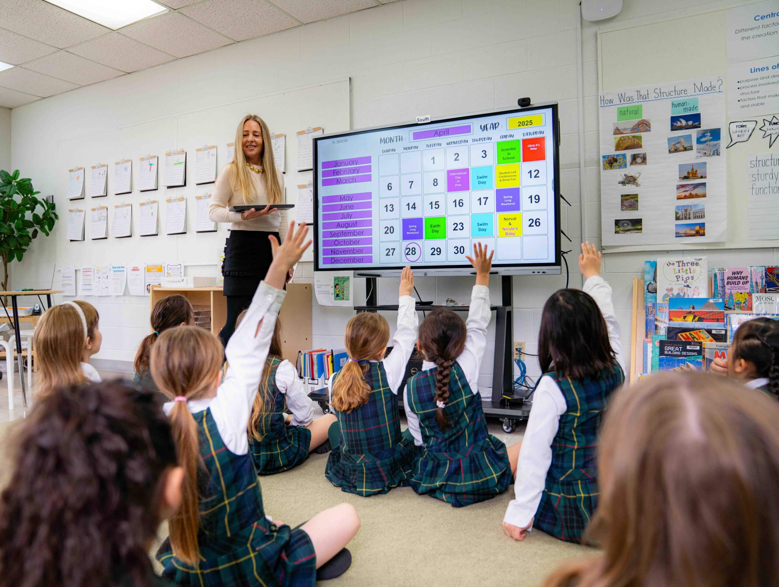 A teacher stands at the front of a classroom holding a tablet, while several young students in plaid uniforms sit on the floor, raising their hands to answer questions on a large interactive screen displaying a colorful quiz.