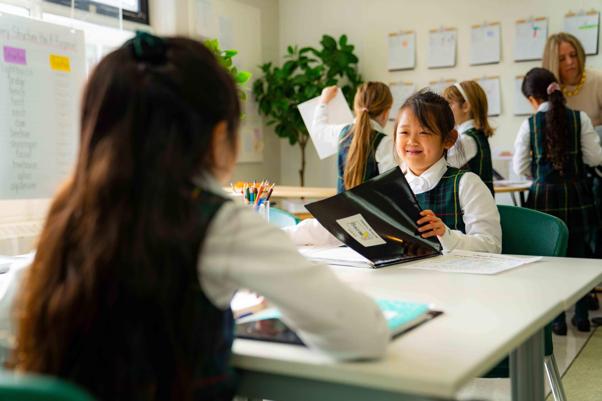 Two young girls in school uniforms sit at a classroom table, smiling and working on assignments with tablets and papers. Other students and a teacher are visible in the background. The room is bright and organized.