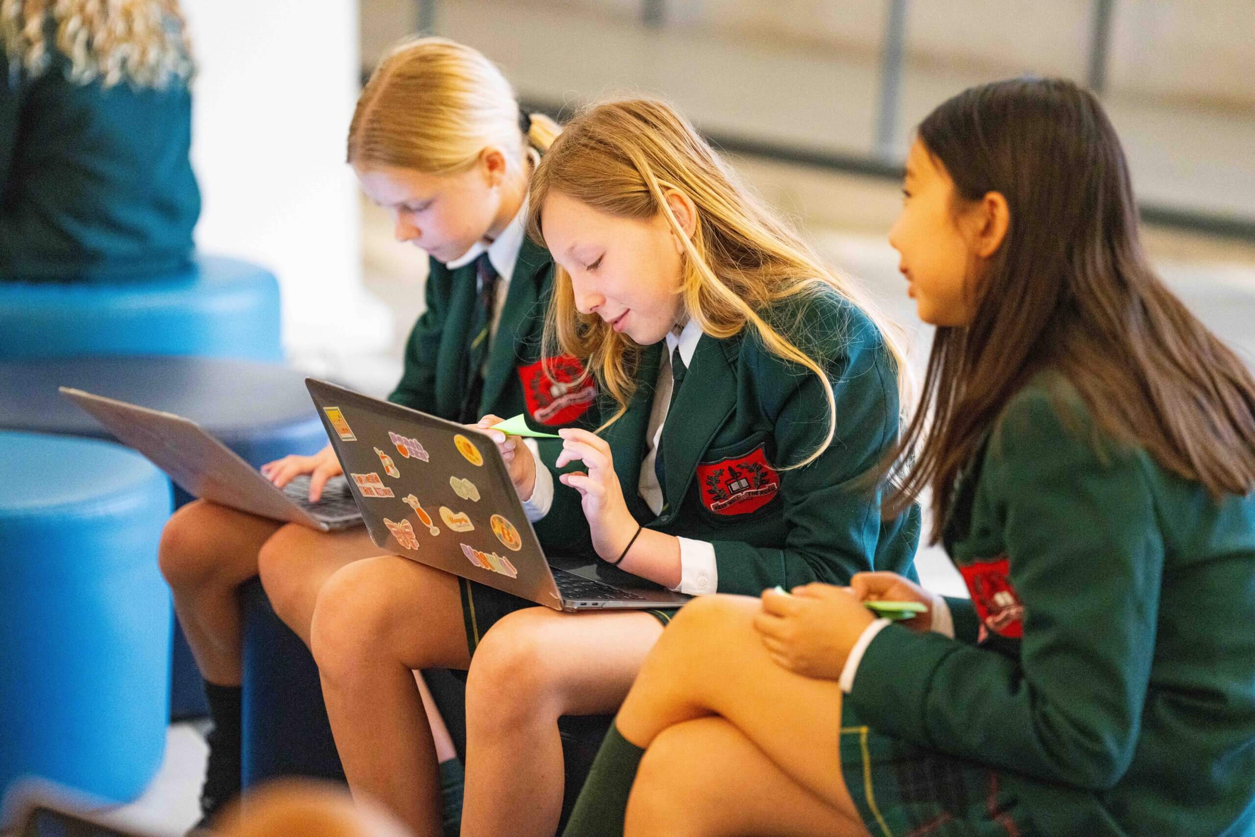 Three girls in green school uniforms sit together, focused on their laptops. Two are typing while the third looks on, smiling, in a brightly lit indoor setting.