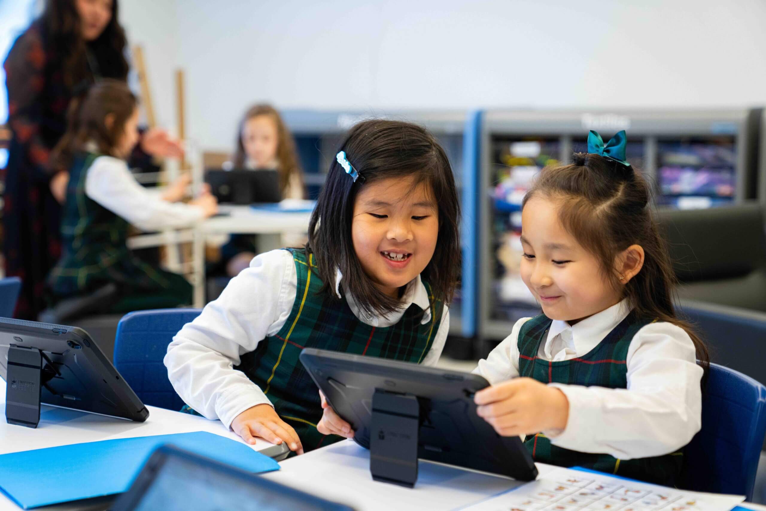 Two young girls in school uniforms smile and look at a tablet together at a classroom table, with other children and a teacher in the background.