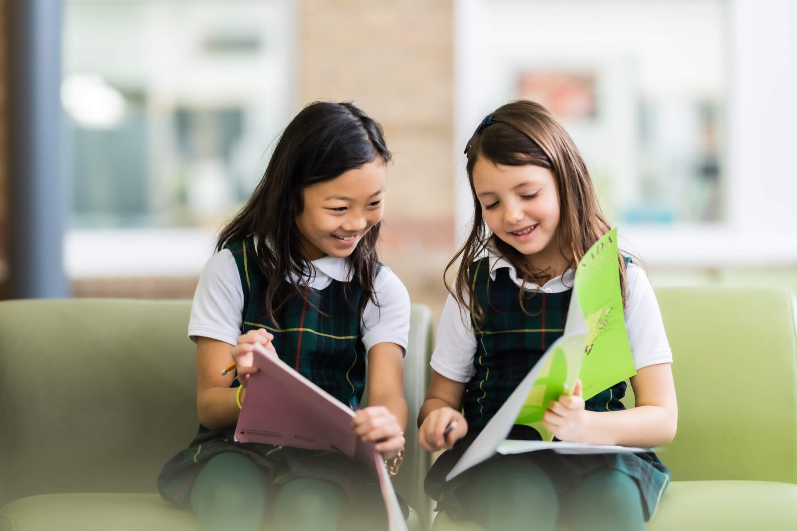 Two young girls in school uniforms sit on a green sofa, smiling and looking at colorful folders or notebooks together, appearing engaged and happy in a bright indoor setting.