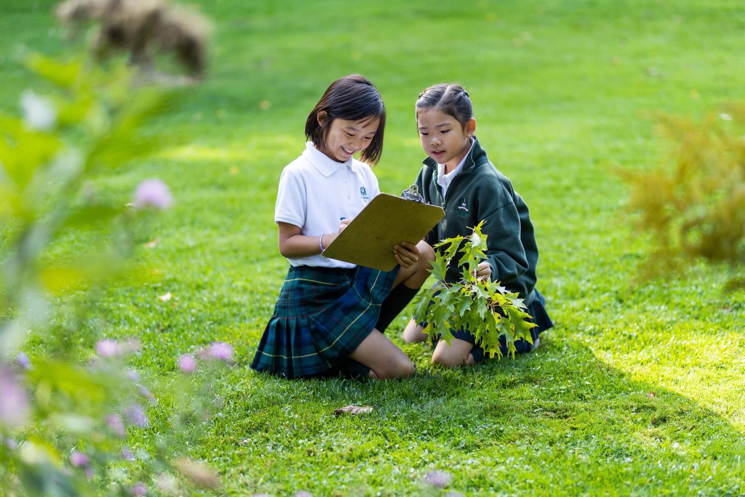 Two girls in school uniforms kneel on grass, studying a clipboard together. One holds a leafy branch, suggesting they are working on a nature project or outdoor assignment in a green park setting.