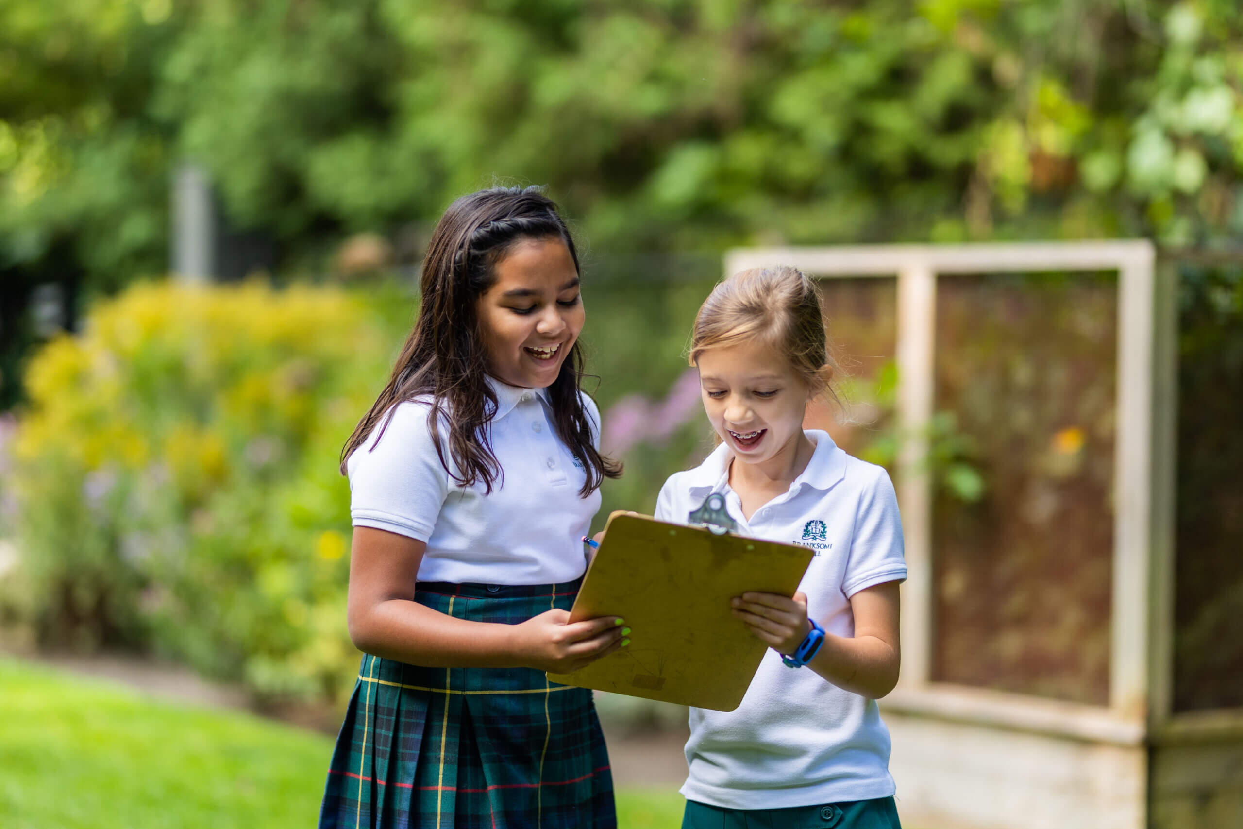 Two young girls in school uniforms smile and look at a clipboard while standing outside in a garden or park, with greenery and trees in the background.
