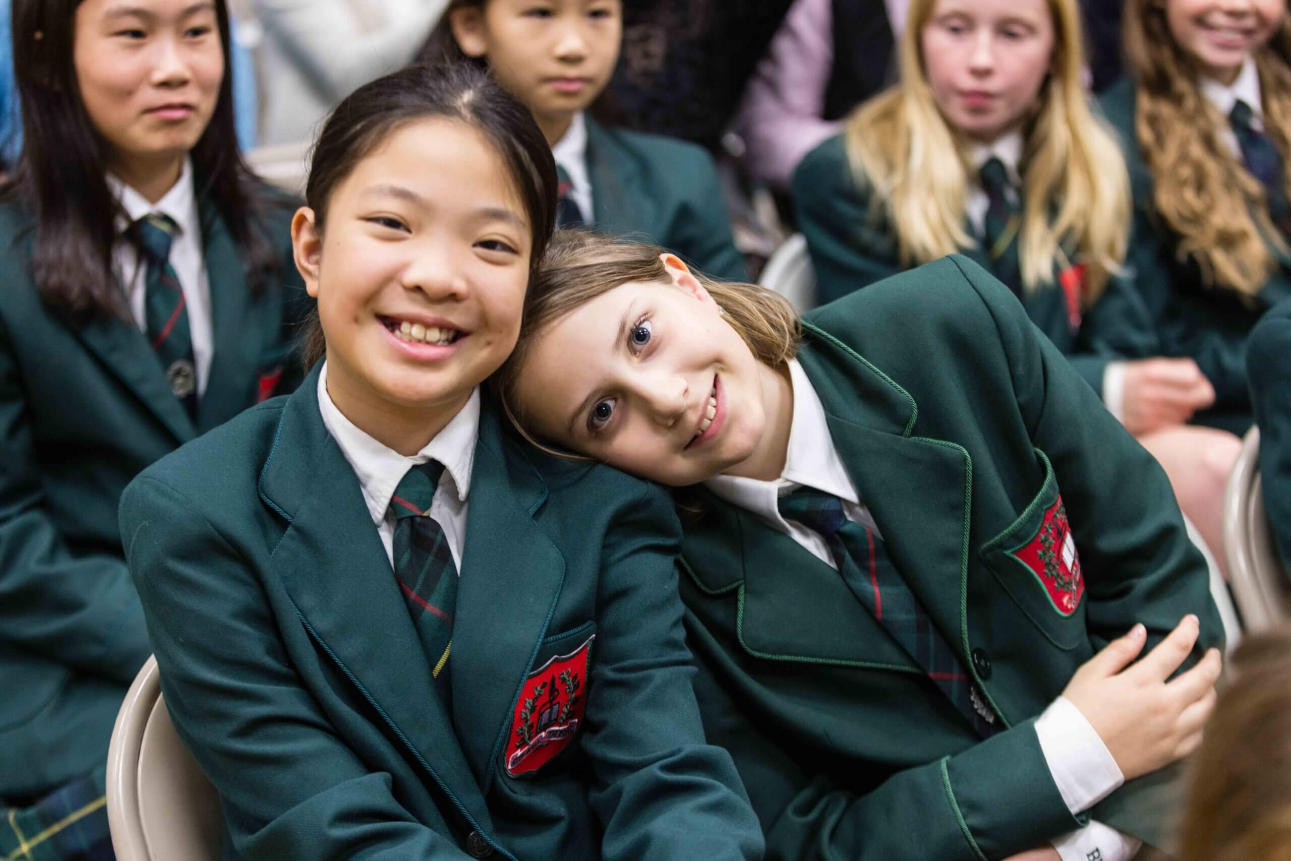 Two students in green blazers, one smiling and the other resting her head on her friend's shoulder, sit together among a group of classmates at what appears to be a school event.
