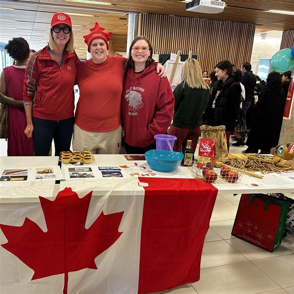 Three people in red and white stand behind a table covered with a Canadian flag, displaying food and items. They are indoors at an event with other tables and attendees in the background.