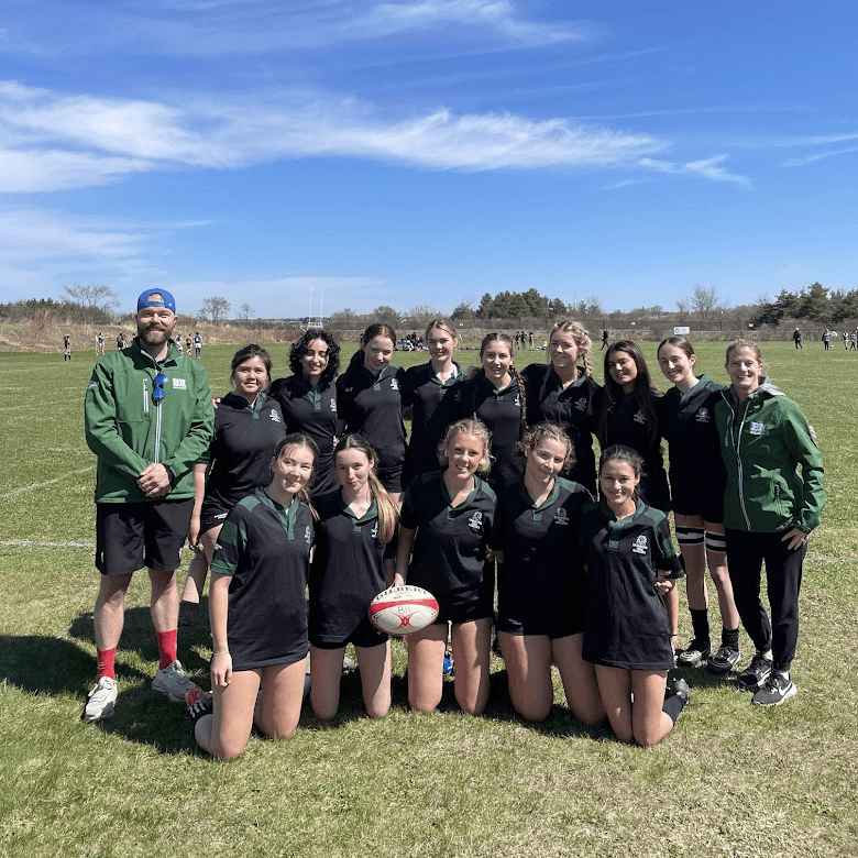 A girls' rugby team poses outdoors on a grassy field with their coach. The team members wear black uniforms and smile at the camera, while the coach wears a green jacket and blue cap. A rugby ball is held in front.