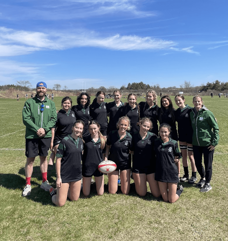 A girls' rugby team poses outdoors on a grassy field with their coach. The team members wear black uniforms and smile at the camera, while the coach wears a green jacket and blue cap. A rugby ball is held in front.