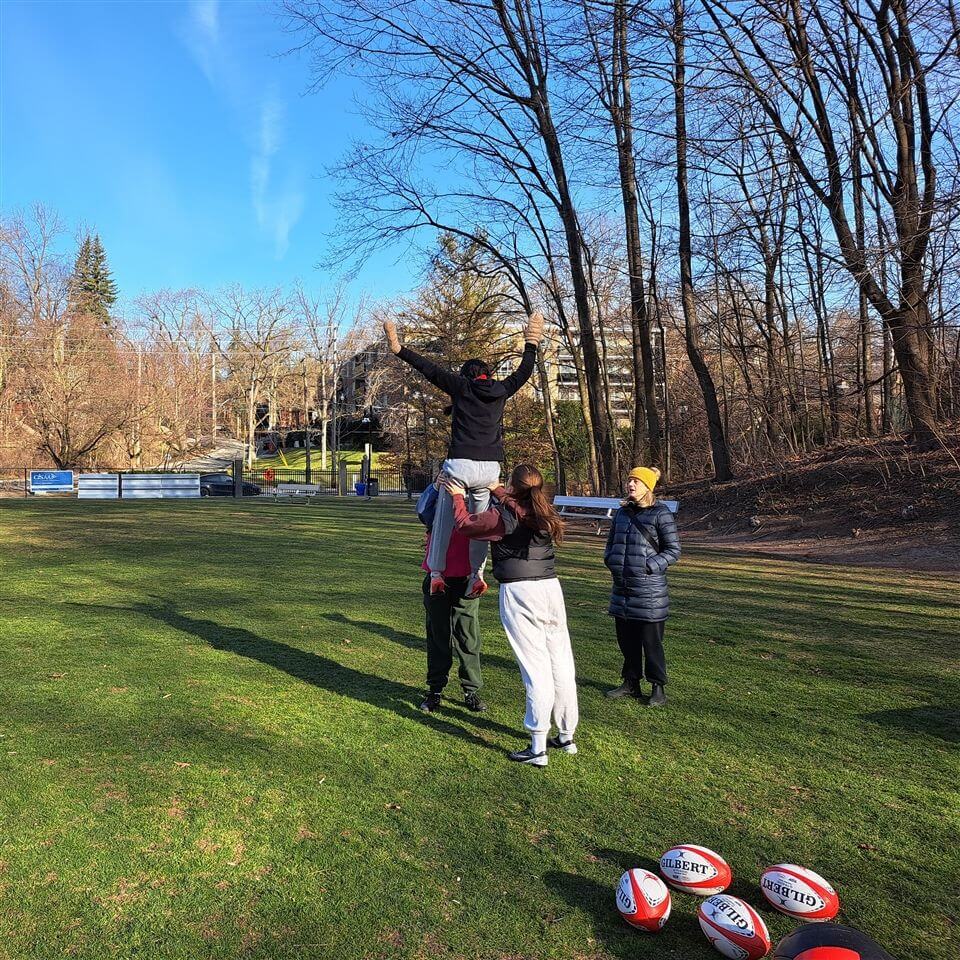Four people are outdoors on a grassy field. Two are lifting a third person into the air, while a fourth looks on. Several red, white, and blue rugby balls are on the ground nearby. Trees and buildings are in the background.