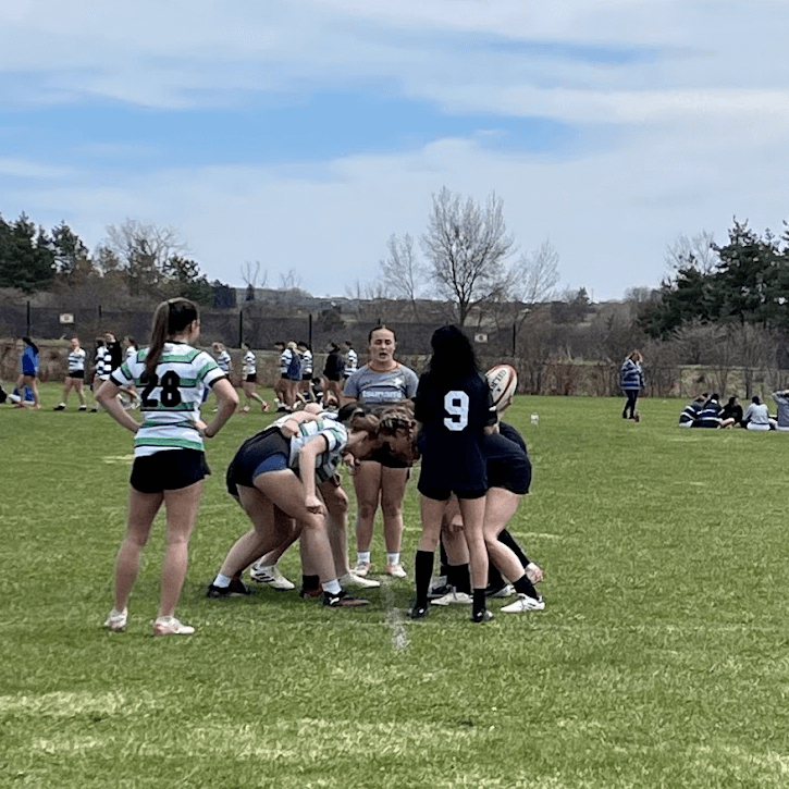 A women's rugby match in progress outdoors; players in striped and dark jerseys are preparing for a scrum on a grassy field with spectators and teams in the background under a cloudy sky.