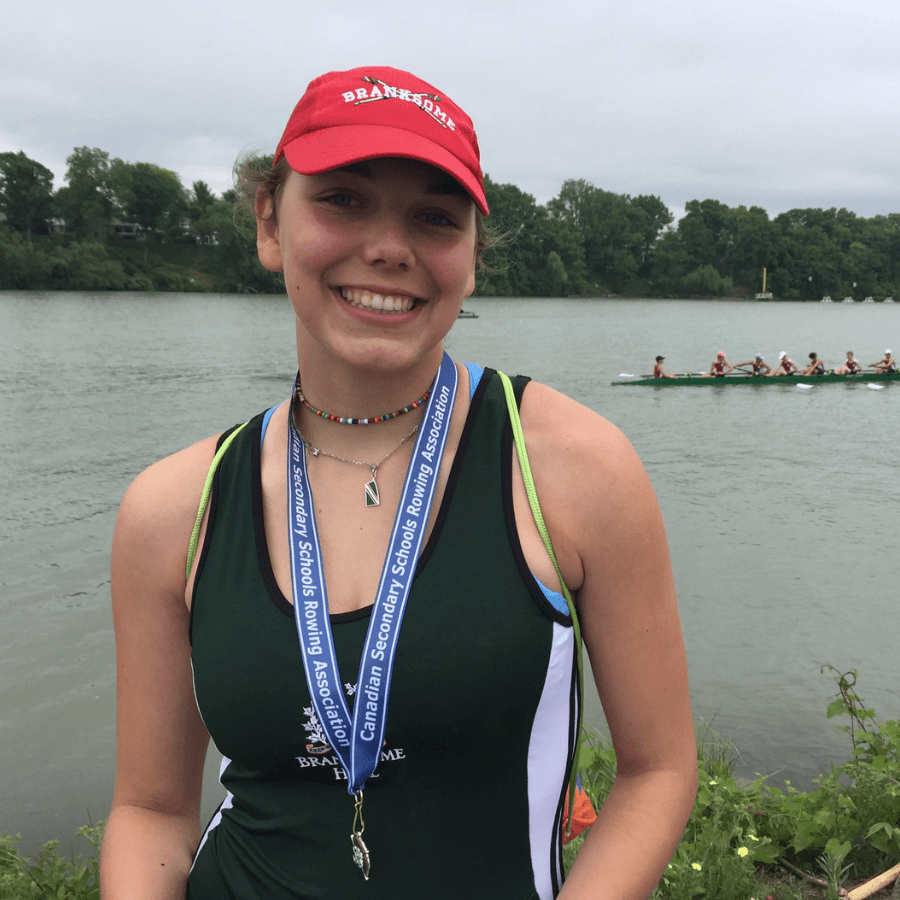 A smiling girl wearing a red “Branksome” cap and a medal stands by a river with a rowing team in a boat in the background. She is dressed in a rowing uniform and greenery is visible along the riverbank.