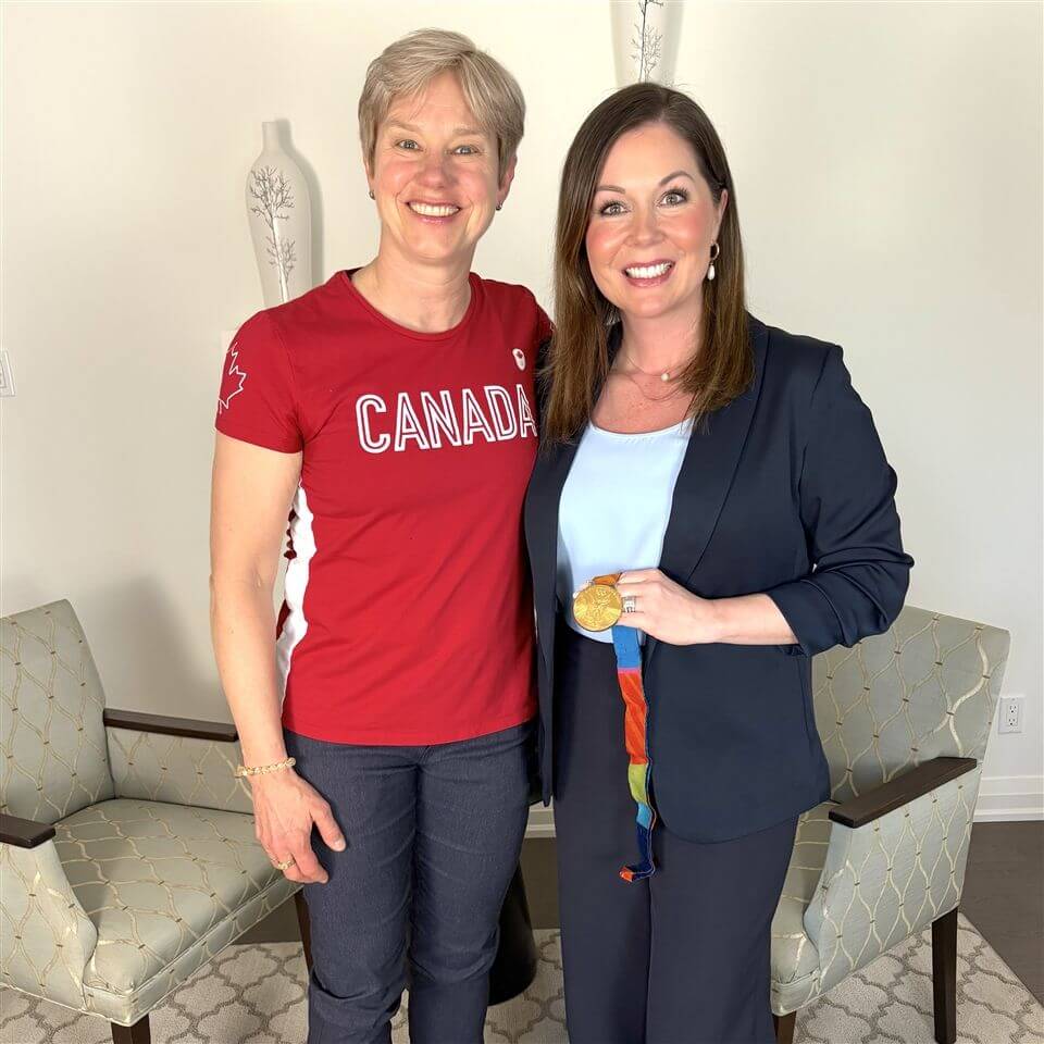 Two women stand and smile for the camera. The woman on the left wears a red "Canada" shirt, and the woman on the right, in a blazer, holds a gold medal with a colorful ribbon. They are indoors with chairs and lamps in the background.