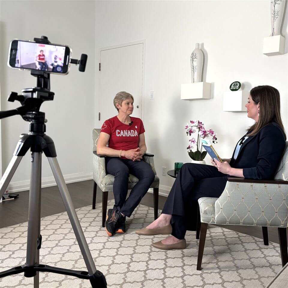 Two women sit facing each other in armchairs during an interview; one wears a red "CANADA" shirt, while the other holds a notepad. A smartphone on a tripod records the conversation in a bright, modern room.