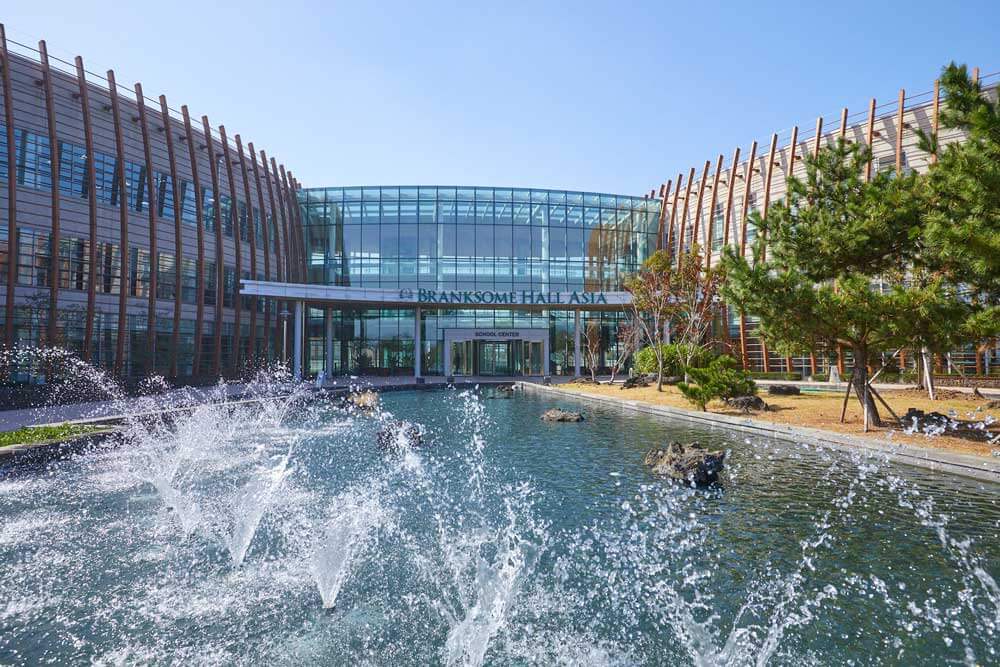 A modern glass building with wooden accents labeled "Branksome Hall Asia" stands behind a rectangular pond with multiple water fountains spraying upwards, surrounded by landscaped trees under a clear blue sky.