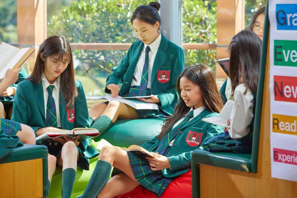 Four students in green school uniforms sit together reading books in a bright, sunny room with large windows and greenery outside. A colorful educational poster is visible on the right.