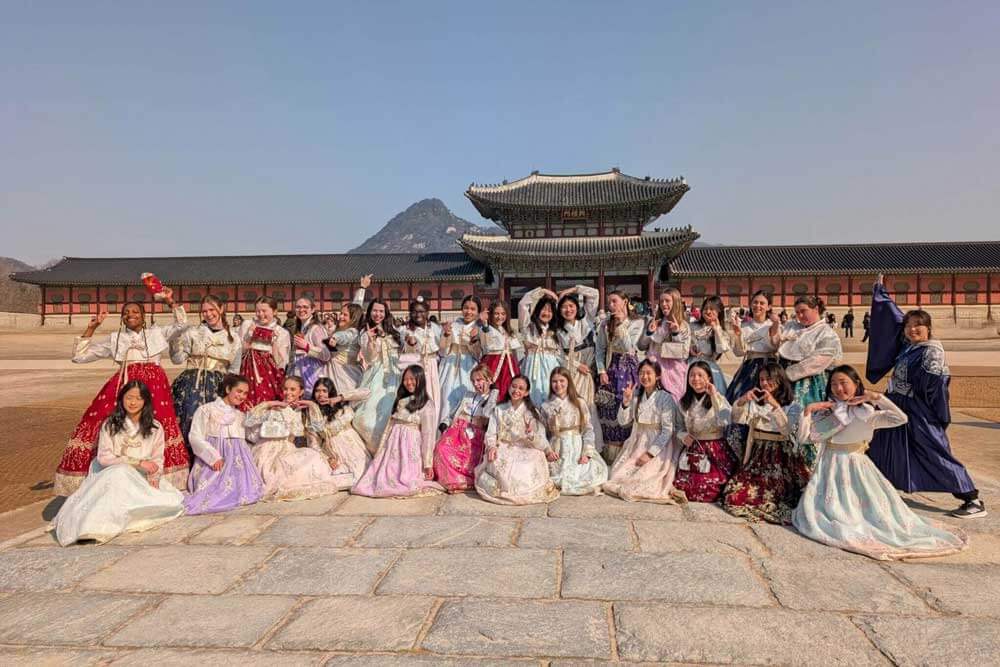 A large group of women pose together wearing colorful traditional hanbok dresses in front of a historic palace building, with a mountain visible in the background under a clear sky.