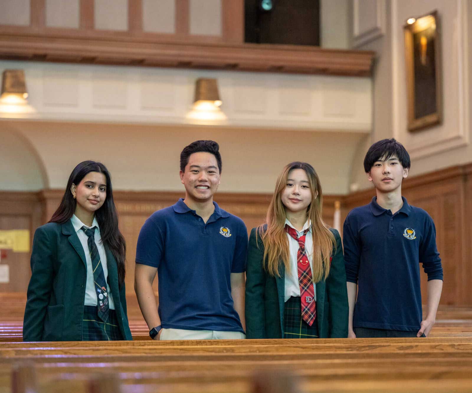 Four students in school uniforms stand inside a church-like hall with wooden pews, smiling and facing the camera. Two wear blazers and ties, while two wear blue polo shirts with the school's emblem.