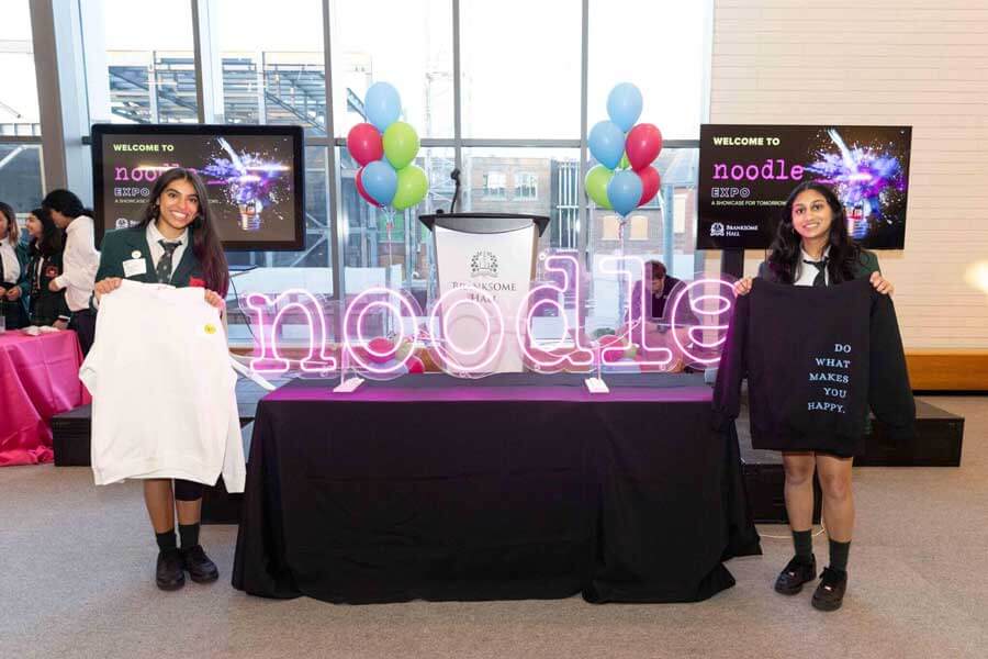 Two women stand on either side of a table with a neon "noodle" sign, holding up shirts. Balloons and screens are in the background, and the event appears to be indoors.
