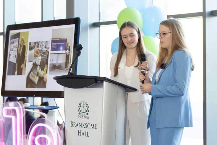 Two young women in business attire stand at a podium labeled "Branksome Hall," speaking into microphones. Behind them is a screen displaying scientific or medical images and blue and green balloons.