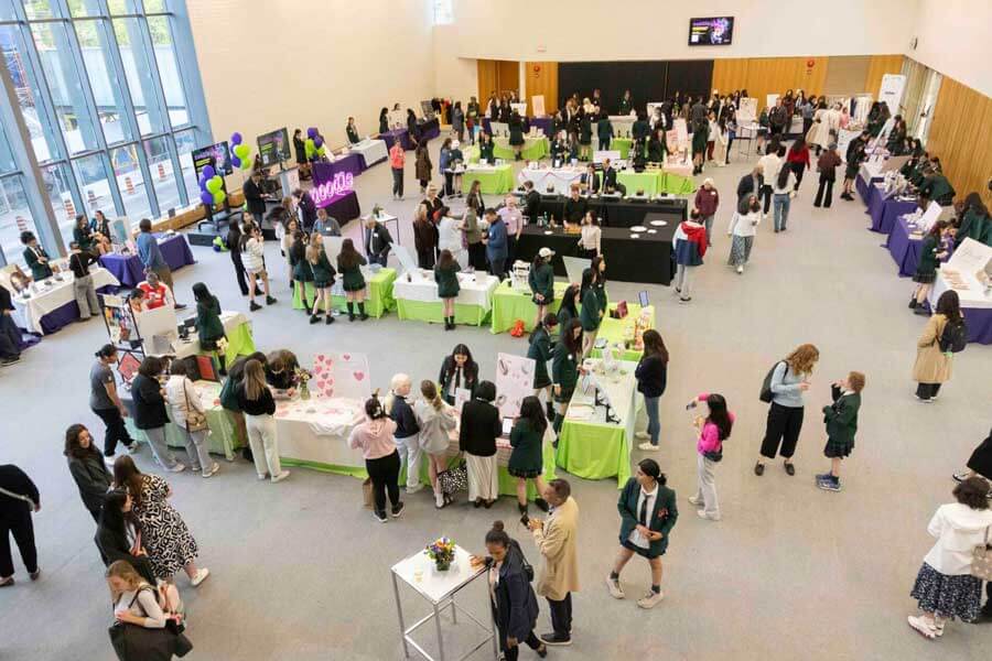 A large room filled with people attending an indoor event with tables covered in colorful tablecloths, displays, and informational booths. Attendees interact around the exhibits and walk through the open space.