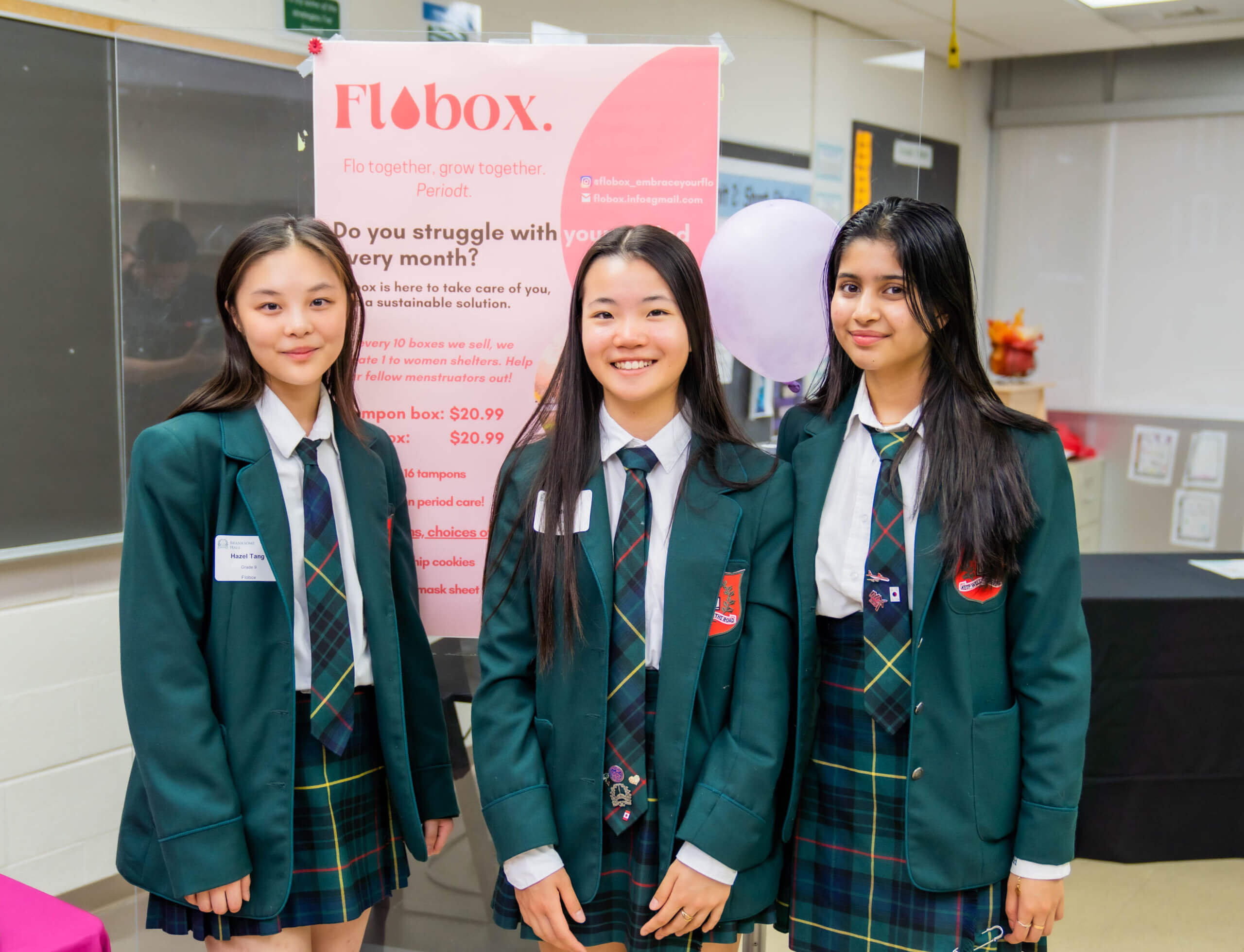 Three high school students in green uniforms stand in front of a Flobox display board at a school event. They are smiling and posing for the photo. The background shows classroom elements and posters.