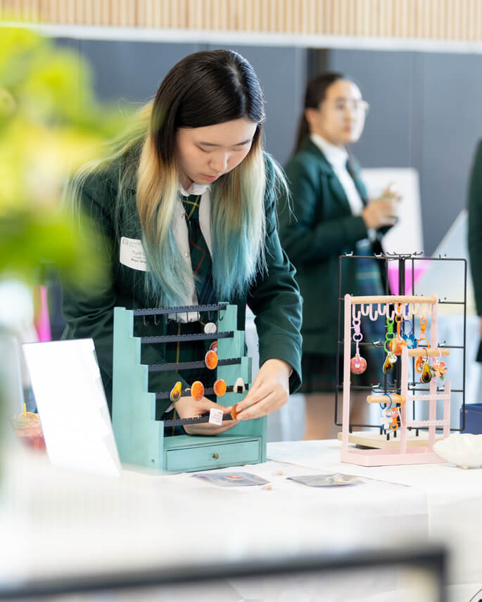 A student in a green uniform arranges colorful handmade jewelry on a display stand at a table during a school event. Another student in a similar uniform stands in the background.