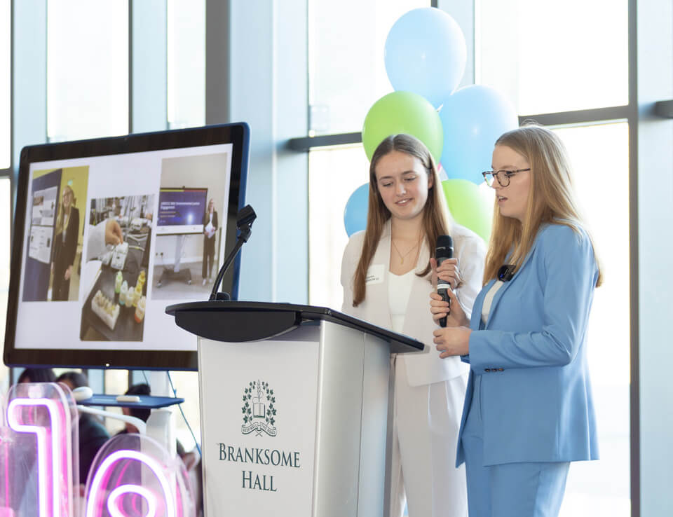 Two young women in business attire speak at a podium labeled "Branksome Hall." One holds a microphone while the other stands beside her. Behind them, a screen displays images and there are blue and green balloons.