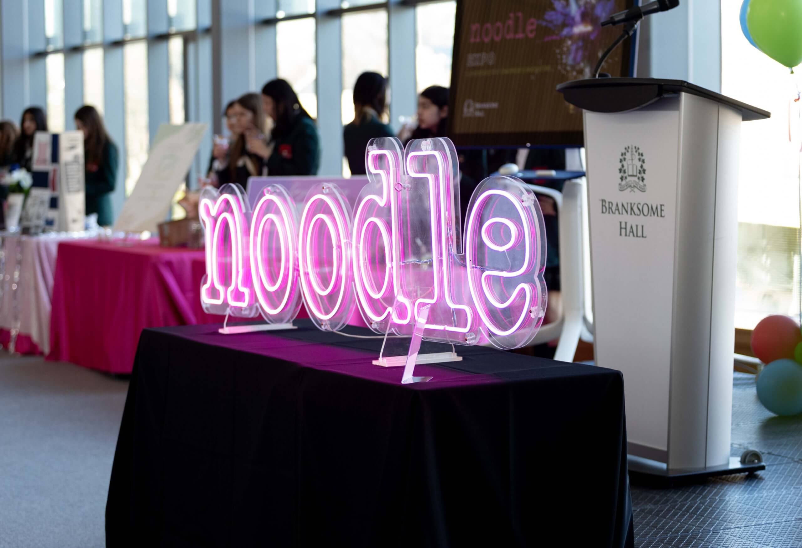 A neon pink "noodle" sign sits on a black tablecloth in a bright room. In the background are people, booths, and a podium labeled "BRANKSOME HALL.