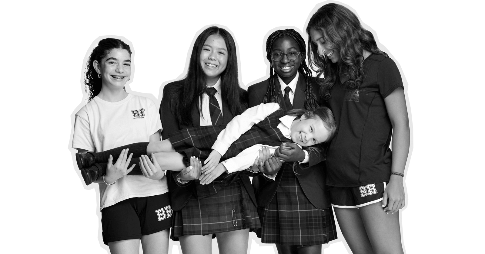 Five girls in school uniforms and sportswear stand together smiling. Two of them hold a younger girl horizontally across their arms. They all appear happy and friendly, posing for a group photo against a plain background.