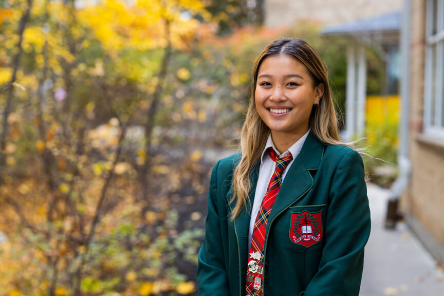 A young woman in a green school blazer and plaid tie stands outside, smiling, with autumn foliage and a school building in the background.