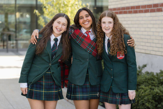 Three smiling girls in green school uniforms with plaid skirts stand together outside, arms around each other, in front of a building with glass windows and brick siding.