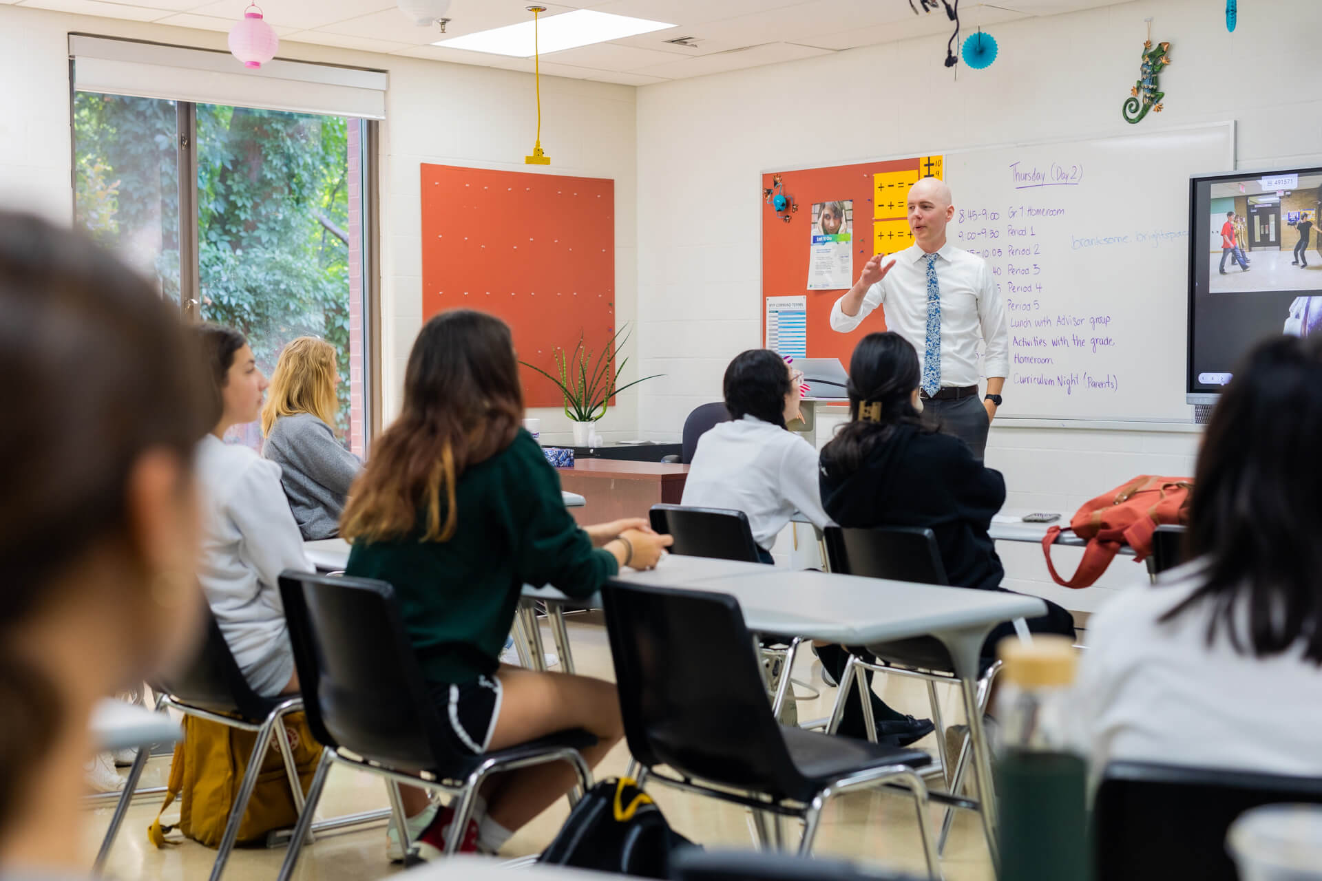 A teacher stands at the front of a classroom, speaking to students seated at desks. The classroom has white walls, bulletin boards, and a whiteboard with notes written on it. The students face the teacher attentively.