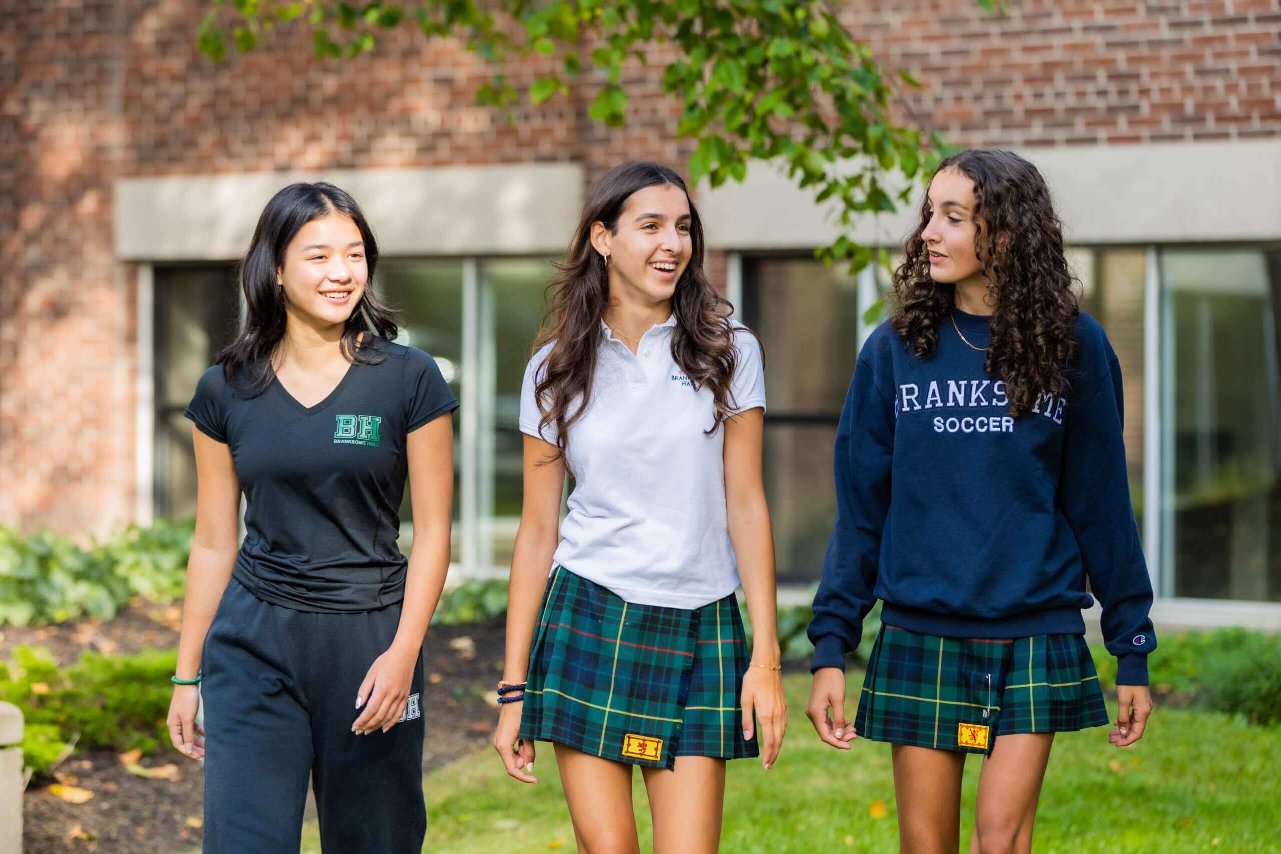 Three teenage girls in school uniforms walk and talk outside near a brick building. Two wear plaid skirts and sweatshirts, and one wears black pants and a black t-shirt. Green grass and trees are in the background.