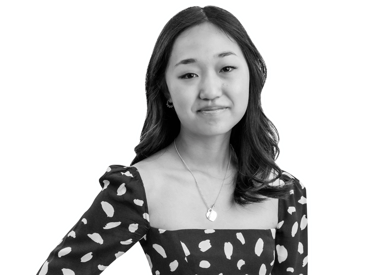 A young woman with long hair, wearing a square-neck dress with a bold dotted pattern and a heart necklace, stands smiling gently against a plain background.
