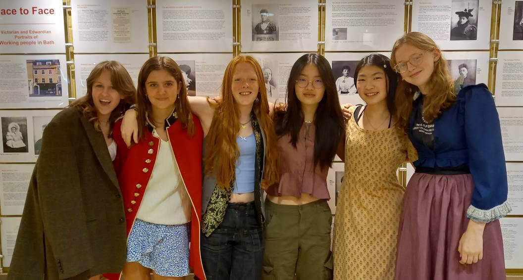 Six smiling teenagers stand arm-in-arm in front of an exhibit with historical photos and texts. They wear colorful and varied outfits, suggesting a casual or celebratory event in a museum or educational setting.