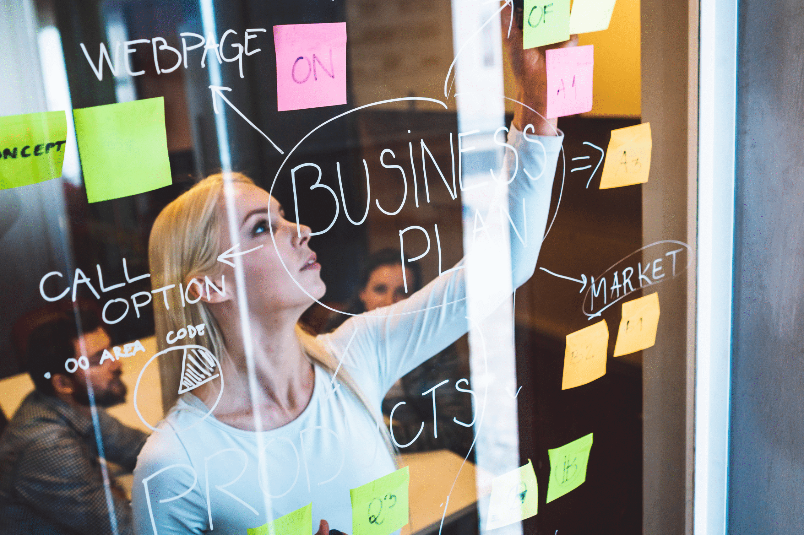 A woman writes on a glass wall covered with sticky notes and diagrams labeled “Business Plan”, while two people sit and discuss behind her in a modern office setting.