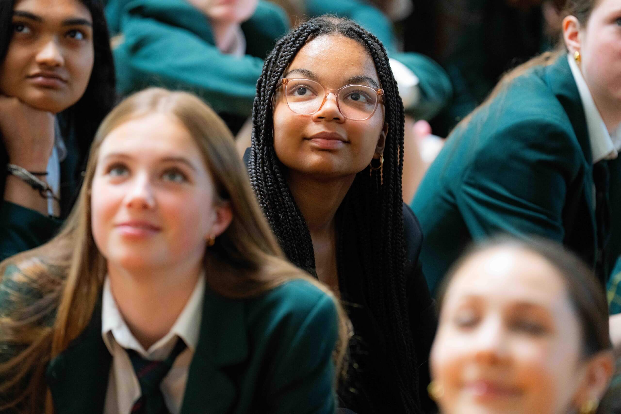 A group of students in green uniforms sit closely together, listening attentively. The focus is on a girl with braids and glasses in the center, looking engaged. Other students around her appear interested and attentive.