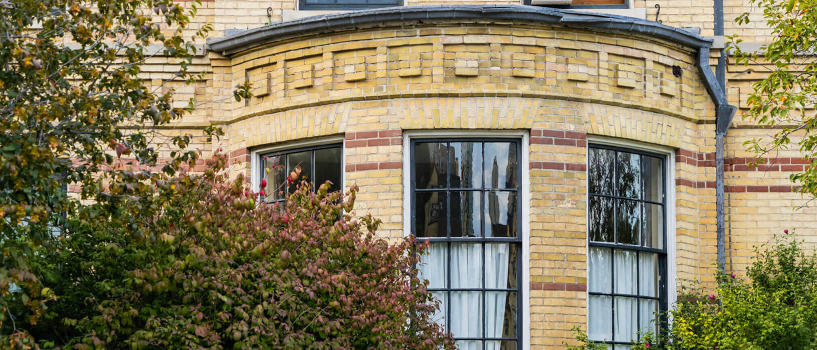 Close-up of a yellow brick building with two large, multi-panel bay windows. Red brick accents run horizontally between the windows. Green shrubs and trees partially obscure the lower part of the building.