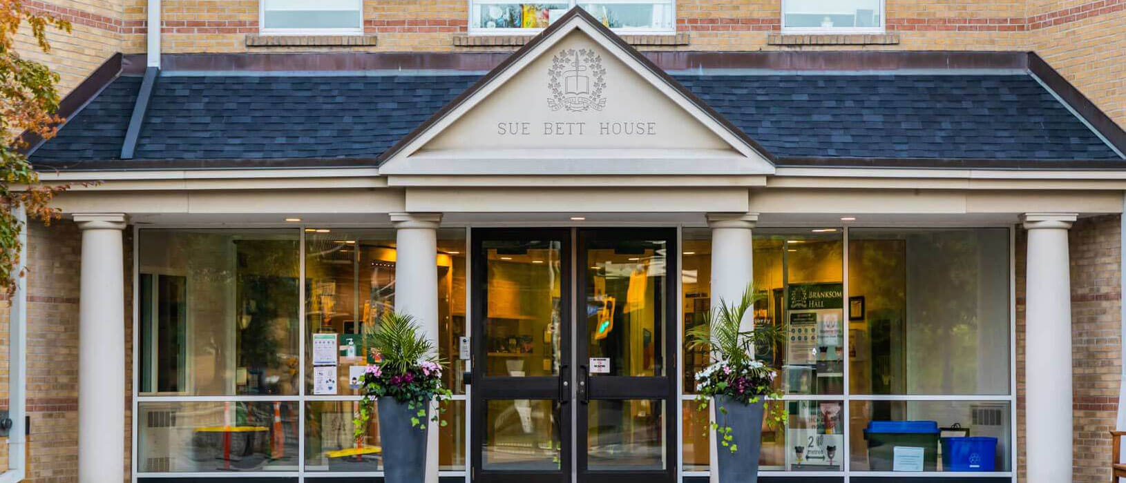 Front entrance of Sue Bett House, a brick building with a triangular pediment, columns, glass doors, and large potted plants on either side of the doorway. The building's name is engraved above the entrance.