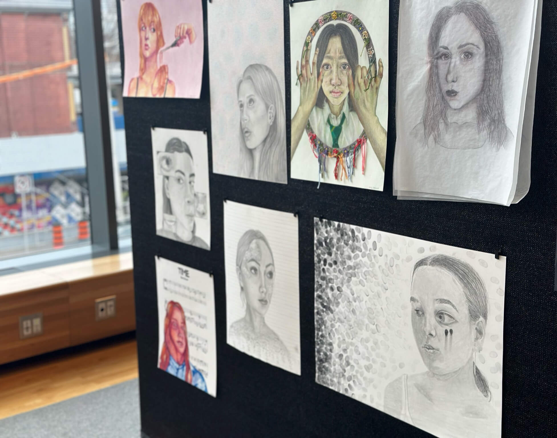 A display board shows several hand-drawn portraits, mostly of women, created with pencil and colored pencil. The board is set up near large windows overlooking a city street.