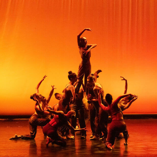 A group of dancers performs on stage with one dancer elevated above the rest, striking an elegant pose. The background is illuminated with a warm orange light, creating a dramatic and artistic atmosphere.