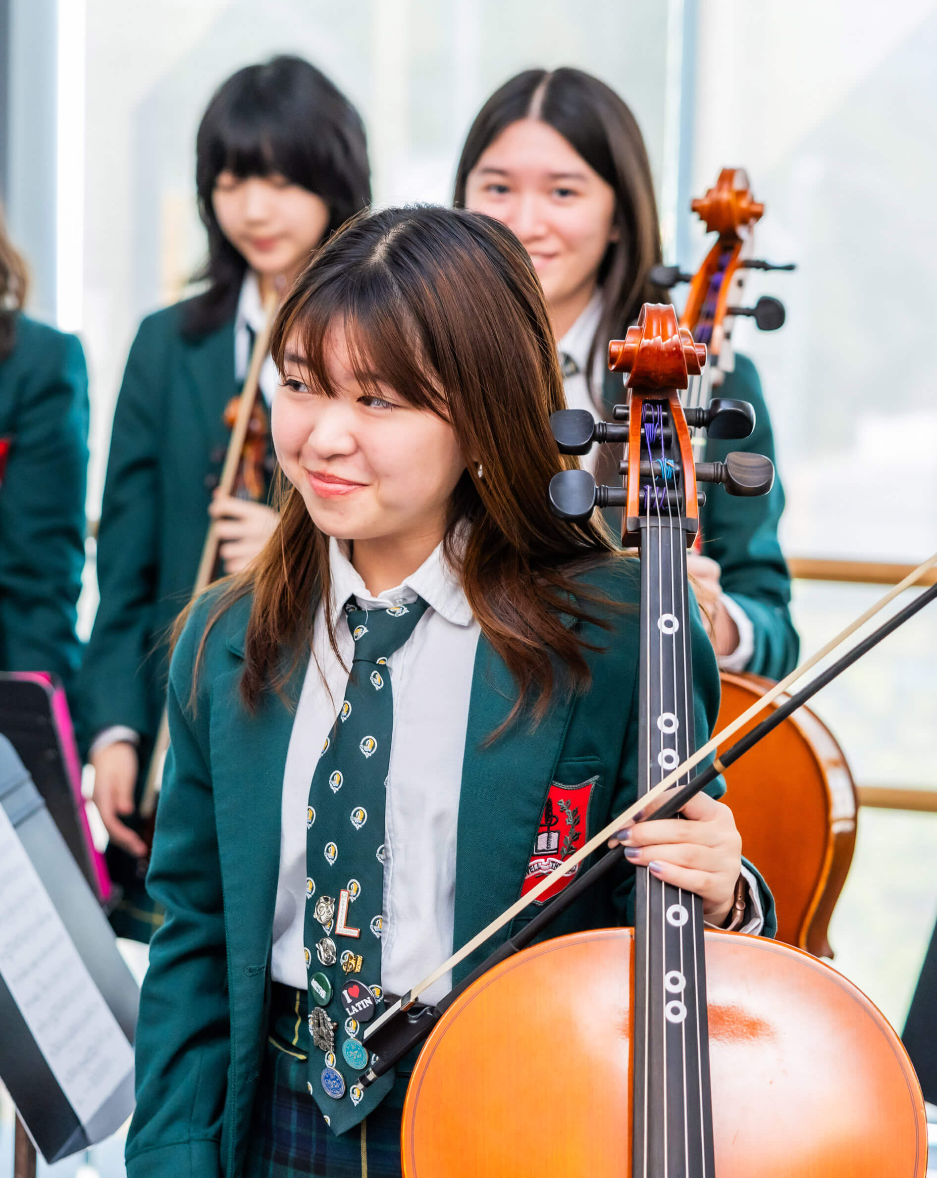 A young woman in a green blazer and tie stands holding a cello, smiling, with other students and string instruments in the background, inside a bright room.