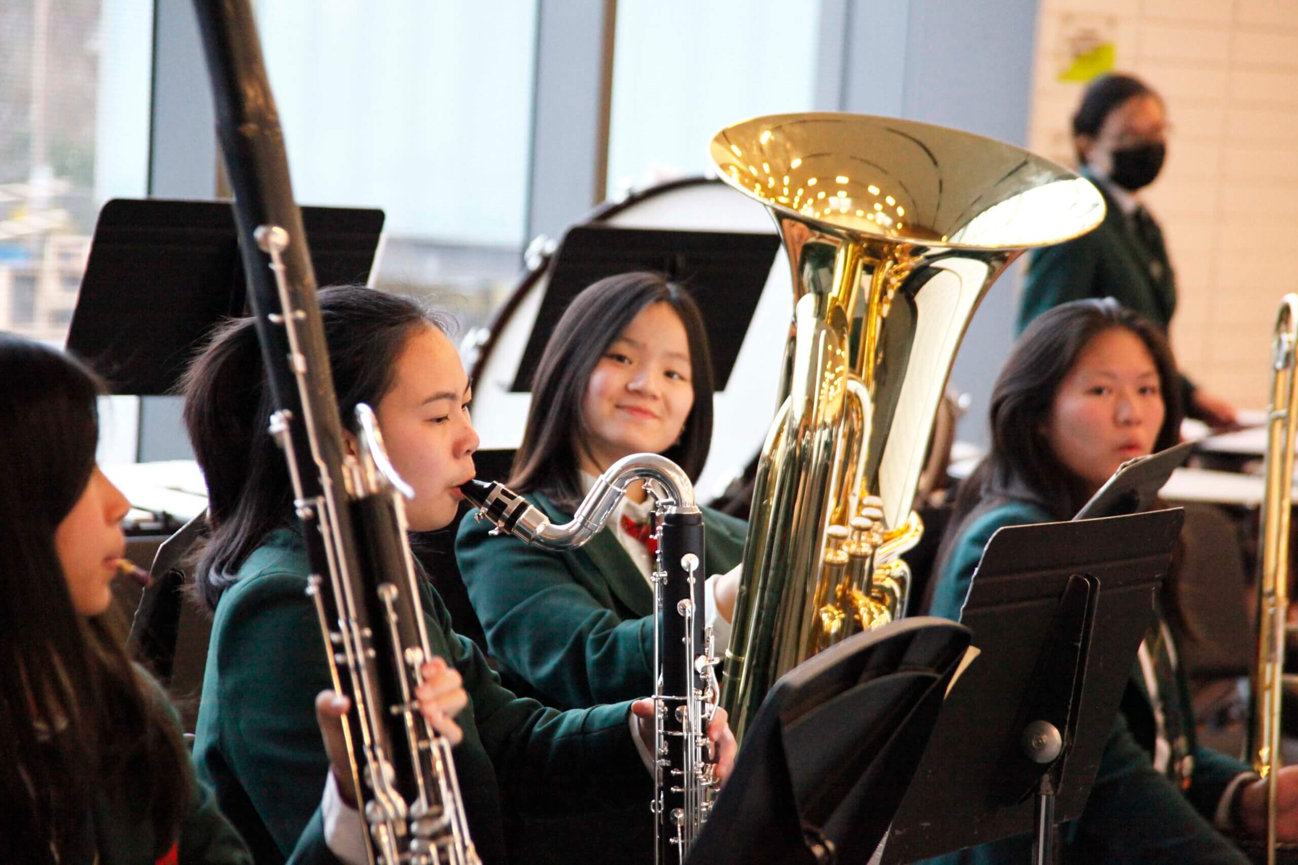 A group of students in green uniforms play wind instruments, including a bassoon and tuba, during a band rehearsal in a brightly lit room. One student in the background wears a black face mask.