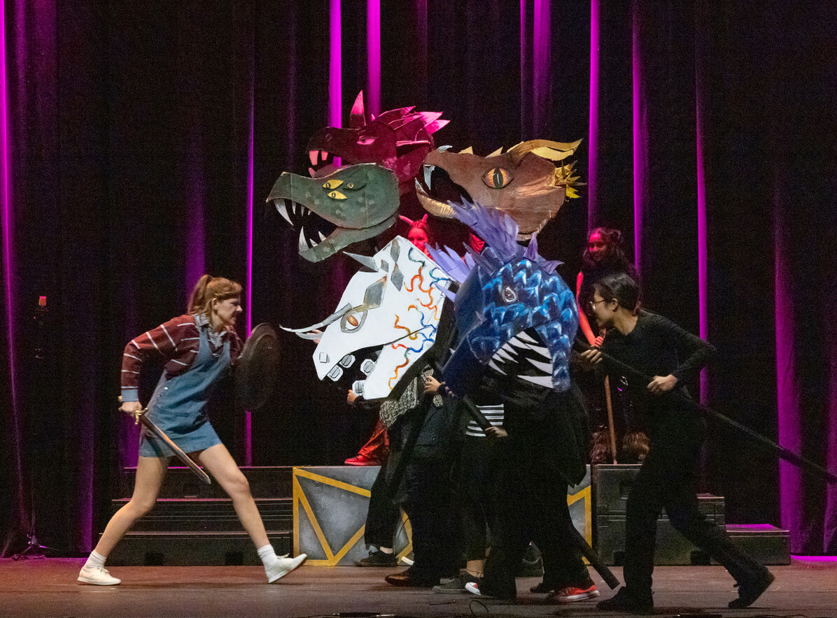 A girl in overalls and a striped shirt faces a group of people dressed in black who are holding large, colorful dragon head puppets on a stage with purple lighting.