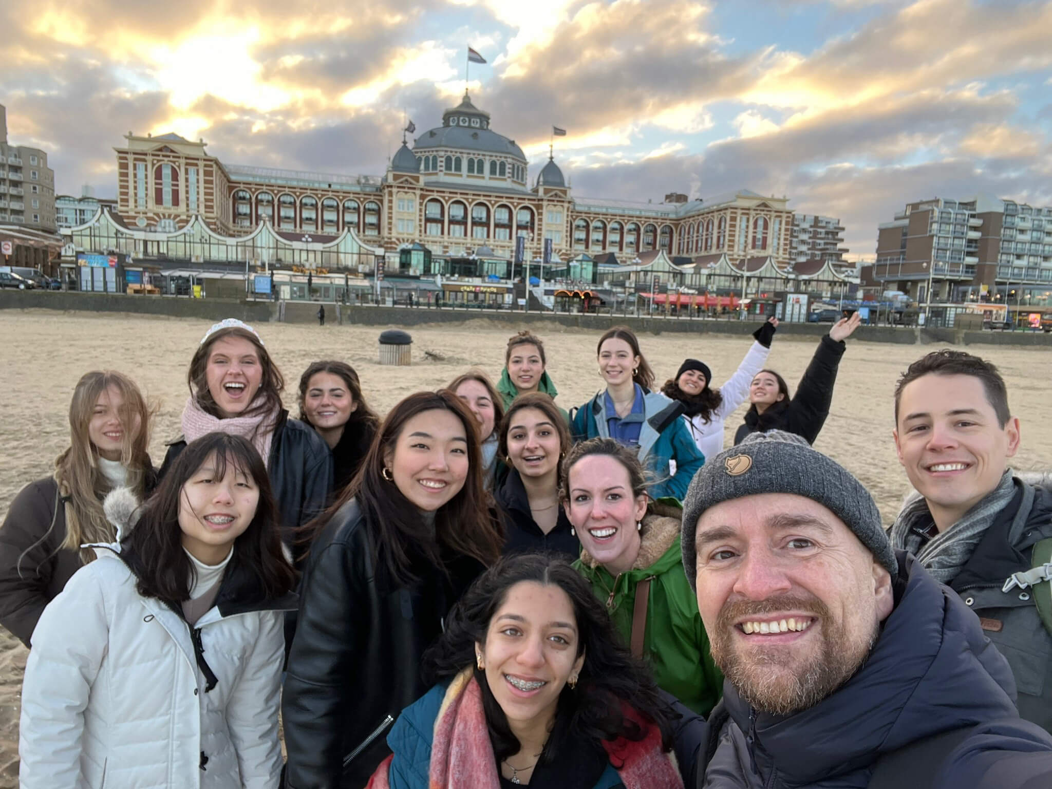 A group of smiling people take a selfie on a sandy beach, with a large, ornate building and cityscape in the background under a cloudy sky at sunset.
